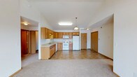 This single-floor space features an open-concept layout that seamlessly connects the kitchen and living area, creating a spacious, functional environment. The kitchen is outfitted with honey-toned oak cabinetry offering generous storage across both upper and lower cabinets. White appliances, including a refrigerator, range, and microwave, are neatly integrated into the layout. A peninsula with additional cabinetry and a built-in sink extends into the open area, providing extra prep space and a natural divide between the kitchen and living zones. Overhead, a flush fluorescent light fixture and a pendant light illuminate the kitchen, while a second pendant adds warmth near the transition to the living area. The kitchen features neutral tile flooring, which gives way to plush carpet in the living area, adding comfort underfoot. The ceilings show subtle variation in height, contributing to the sense of openness. Two interior doors are visible — one leading to additional rooms and another likely serving as an entry or utility access — both featuring classic raised-panel detailing. The overall palette is light and neutral, giving the space a clean, welcoming character.