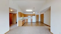 This single-floor space features an open-concept layout that seamlessly connects the kitchen and living area, creating a spacious, functional environment. The kitchen is outfitted with honey-toned oak cabinetry offering generous storage across both upper and lower cabinets. White appliances, including a refrigerator, range, and microwave, are neatly integrated into the layout. A peninsula with additional cabinetry and a built-in sink extends into the open area, providing extra prep space and a natural divide between the kitchen and living zones. Overhead, a flush fluorescent light fixture and a pendant light illuminate the kitchen, while a second pendant adds warmth near the transition to the living area. The kitchen features neutral tile flooring, which gives way to plush carpet in the living area, adding comfort underfoot. The ceilings show subtle variation in height, contributing to the sense of openness. Two interior doors are visible — one leading to additional rooms and another likely serving as an entry or utility access — both featuring classic raised-panel detailing. The overall palette is light and neutral, giving the space a clean, welcoming character.