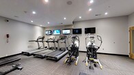 Located at 415-425 N Front St in Columbus, Ohio, this fitness center offers a well-appointed workout facility designed to support an active lifestyle. The space features a clean, modern aesthetic with neutral gray walls, recessed lighting, and patterned carpet flooring that provides both comfort and durability underfoot. The cardio area is equipped with multiple treadmills and elliptical cross-trainers, arranged to maximize the available floor space while maintaining an open and accessible layout. A dedicated wall-mounted air conditioning unit ensures a comfortable environment during workouts regardless of the season. Entertainment is well covered, with several flat-screen TVs mounted at eye level across the walls, allowing users to catch up on news, sports, or shows while exercising. The bright, evenly distributed recessed lighting gives the room a clean and energetic atmosphere. The overall design of the fitness center is practical and functional, offering everything needed for a focused cardio workout in a tidy, well-maintained environment.