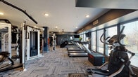 Located at 1330 Kelly Johnson Blvd in Colorado Springs, CO, this well-appointed fitness center offers a comprehensive workout environment designed to support a wide range of exercise routines. The space is finished with a contemporary aesthetic, featuring deep blue-gray walls, recessed lighting, and durable patterned carpet tiles throughout, creating a sleek and functional atmosphere.

The cardio zone is lined with multiple treadmills and elliptical machines positioned along a wall of large windows, allowing natural light to fill the space while providing views of the surrounding Colorado landscape. A wall-mounted television accompanies the cardio area for entertainment during workouts.

The strength training section is generously equipped with a cable crossover machine, a multi-function cable station, a punching bag, weight benches, and additional resistance machines, providing ample options for full-body strength training. Resistance bands and a variety of exercise balls are also available for functional fitness and flexibility work.

A separate studio area, accessible through a glass-paneled door, features windowed pass-throughs and its own dedicated space for group or individual training. A small refreshment station with cabinetry is conveniently located near the studio entrance. The overall layout is open and spacious, offering room to move freely between equipment zones.