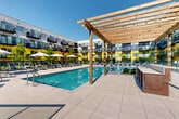 Located along Philadelphia's waterfront at 501 North Christopher Columbus Boulevard, this outdoor amenity space is a thoughtfully designed retreat that blends modern architecture with lush, natural landscaping. The centerpiece is a large resort-style swimming pool, surrounded by an expansive concrete deck outfitted with lounge chairs and market umbrellas, all shaded by a striking wood pergola structure. Adjacent to the pool, a lively courtyard features raised planters filled with native grasses, flowering shrubs, and ornamental greenery, creating a vibrant and inviting atmosphere throughout the warmer months. Broad, light-toned paved pathways connect multiple outdoor zones, including a turf lawn area lined with additional lounge seating — the perfect spot for sunbathing or relaxing in the open air. Glass panel railings frame a dedicated waterfront terrace with composite wood decking, offering unobstructed views of the Delaware River and the iconic Benjamin Franklin Bridge. The surrounding multi-story building features a bold, contemporary façade with vivid yellow and gray paneling, private balconies, and large windows. The entire space is bathed in natural light, benefiting from its open-air design and prime riverfront orientation.