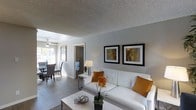 This single-floor residence offers a thoughtfully designed open-concept layout that connects the living, dining, and bedroom areas in a smooth, functional flow. The living room serves as the heart of the space, featuring light grey walls, wood-style flooring, and a textured ceiling that adds subtle depth overhead. A white tufted sofa anchors the seating area, accented by warm amber throw pillows and complemented by a sleek coffee table and side lamp, creating an inviting and contemporary atmosphere. Two framed wall art pieces add a curated touch to the main wall. Natural light filters in through slatted blinds on the windows, brightening the interior throughout the day. A ceiling fan is visible in the adjacent dining area, which features a dark dining set that contrasts elegantly with the lighter surroundings. The open sightlines between rooms enhance the sense of spaciousness throughout the unit. A hallway leads to the private bedroom and additional rooms beyond. The overall aesthetic is clean, modern, and cohesive, with a neutral color palette punctuated by warm accent tones, making this a comfortable and stylish single-level living space.