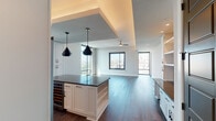 This contemporary single-floor residence showcases a thoughtfully designed open-concept layout that seamlessly connects the kitchen and living areas. The kitchen is a standout feature, equipped with crisp white shaker cabinetry extending to the ceiling, stainless steel appliances including a gas range, built-in microwave, and french door refrigerator, complemented by a large center island with dark countertops and a built-in wine cooler. Geometric black pendant lights hang above the island, and a tile backsplash adds refined texture. Rich, dark hardwood flooring flows throughout the main living areas, while floor-to-ceiling windows flood the space with natural light and offer sweeping city skyline views. A built-in shelving and desk unit provides additional functionality in the living area. The bedrooms feature plush carpeting, ceiling fans, and large windows framing urban vistas. The primary bathroom is generously proportioned, with a dual-sink vanity featuring dark countertops, white cabinetry, a glass-enclosed walk-in shower with tile surround, and elegant pendant lighting. The secondary bathroom mirrors the refined aesthetic with a soaking tub, tiled surround, and stylish pendant light. Throughout, dark-paneled interior doors provide a striking contrast to the light, neutral wall tones.