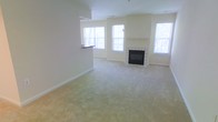 This single-floor space centers around a generously sized living area finished with soft, neutral-toned wall-to-wall carpeting that adds warmth underfoot. The room's most notable feature is a classic white tile-surround fireplace positioned along one wall, flanked by multiple windows that allow abundant natural light to flow throughout. The windows offer views of surrounding greenery, enhancing the airy, open atmosphere of the interior.

The layout includes a partial wall with a raised countertop pass-through, suggesting an adjacent kitchen or dining area that connects seamlessly to the main living space. This architectural detail promotes an open-concept feel while maintaining a subtle sense of separation between functional zones. The walls are finished in a clean, neutral palette that complements the light carpeting and contributes to the overall bright and inviting ambiance.

Elevated ceiling lines add a sense of spaciousness to the room, reinforcing the open and comfortable character of the space. The combination of natural light, soft finishes, and the focal fireplace creates a welcoming environment well-suited for everyday living.