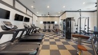 This modern fitness center in Murfreesboro, TN is thoughtfully designed to support a comprehensive workout experience. The space is unified by a bold checkered carpet tile flooring in neutral tones, creating a dynamic visual foundation throughout. High ceilings and recessed lighting give the main workout area an open, energetic ambiance, while ceiling fans provide additional comfort during intense training sessions. The cardio zone is lined with treadmills and ellipticals, each equipped with individual mounted screens, allowing members to stay entertained while training. Strength training is well-supported with a full rack of dumbbells, weight benches, and multiple cable and resistance machines. Large glass garage-style doors open to views of an outdoor patio and pool area, flooding the space with natural light and creating a seamless indoor-outdoor connection. A dedicated spin room houses multiple Spinning bikes alongside medicine balls and hand weights, accented by a textured wood-panel feature wall that adds warmth. The entry area includes a beverage cooler, towel storage, and a small sink for member convenience. Wall-mounted mirrors throughout the strength areas enhance the sense of space and allow users to monitor their form. The overall design is clean, functional, and contemporary.