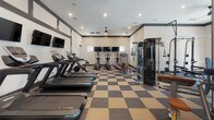 This modern fitness center in Murfreesboro, TN is thoughtfully designed to support a comprehensive workout experience. The space is unified by a bold checkered carpet tile flooring in neutral tones, creating a dynamic visual foundation throughout. High ceilings and recessed lighting give the main workout area an open, energetic ambiance, while ceiling fans provide additional comfort during intense training sessions. The cardio zone is lined with treadmills and ellipticals, each equipped with individual mounted screens, allowing members to stay entertained while training. Strength training is well-supported with a full rack of dumbbells, weight benches, and multiple cable and resistance machines. Large glass garage-style doors open to views of an outdoor patio and pool area, flooding the space with natural light and creating a seamless indoor-outdoor connection. A dedicated spin room houses multiple Spinning bikes alongside medicine balls and hand weights, accented by a textured wood-panel feature wall that adds warmth. The entry area includes a beverage cooler, towel storage, and a small sink for member convenience. Wall-mounted mirrors throughout the strength areas enhance the sense of space and allow users to monitor their form. The overall design is clean, functional, and contemporary.