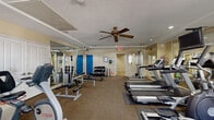 This fitness center offers a comprehensive exercise environment spread across a single floor, thoughtfully organized to accommodate a wide range of workout needs. The main exercise area is generously proportioned, with neutral beige walls, carpet flooring, and ceiling fans providing air circulation throughout the space. Recessed lighting and natural light from multiple windows fitted with blinds contribute to a bright, functional atmosphere.

The cardio section features a row of treadmills, elliptical machines, and stationary bikes, including a recumbent model, catering to varying fitness levels. On the strength training side, a multi-station cable machine, weight bench, and a well-stocked dumbbell rack provide ample options for resistance workouts. Large wall mirrors line portions of the space, enhancing the sense of openness and allowing users to monitor their form.

A flat-screen television is mounted for entertainment during workouts. The facility also includes dedicated restroom facilities, finished with white tile flooring, tiled wainscoting, standard stall partitions, and a shower area, ensuring convenience for all users. The overall layout is practical and accessible, designed to support a complete fitness routine.