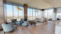This sophisticated high-rise lounge occupies a single expansive floor, defined by dramatic floor-to-ceiling curtain-wall glazing that wraps around the perimeter, delivering panoramic city views from every vantage point. The soaring ceiling height amplifies the sense of openness, while wide-plank light oak flooring grounds the space with warmth and continuity throughout.

The layout is organized into several distinct yet connected seating zones. One area features a set of quilted velvet barrel chairs arranged around a round black side table, anchored by a cowhide rug. Adjacent to this, a larger lounge zone pairs a neutral linen sofa with mid-century-inspired accent chairs and a rectangular coffee table. A smaller intimate seating nook offers an additional sofa and lounge chair positioned directly against the glass to maximize the views. Sculptural cluster pendant lights illuminate each zone, adding a contemporary design element overhead.

A separate dining area includes a round pedestal table surrounded by plush upholstered chairs, positioned in front of floor-to-ceiling windows overlooking the city skyline. Dark wood bookshelves provide functional storage nearby.

The kitchenette features dark cabinetry, a white stone countertop, stainless steel sink, and a textured tile backsplash wall mounted with flat-screen televisions, making it well-suited for events and gatherings.