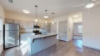 Located at 735W W 15th St in Fayetteville, Arkansas, this single-floor unit offers a thoughtfully designed, modern interior with a cohesive neutral color palette throughout. The open-concept kitchen serves as the centerpiece of the living space, anchored by a large island with granite countertops, an integrated sink, and seating for casual dining. Elegant pendant lighting hangs above the island, complementing the flush-mount ceiling fixtures. The kitchen is equipped with stainless steel appliances — including a refrigerator, electric range, and built-in microwave — and features generous grey cabinetry that provides ample storage. Wood-look plank flooring extends throughout the main living area, lending warmth and a clean, contemporary aesthetic. The bathroom mirrors the kitchen's design language, showcasing matching grey cabinetry, granite countertops, a well-lit vanity mirror, and the same wood-look flooring. The bedroom offers plush carpeting underfoot, a ceiling fan with light fixture, and a window dressed with blinds that allows for natural light. The unit's clean lines, consistent finishes, and functional layout make it a well-appointed and move-in-ready space in the heart of Fayetteville.