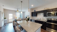 This single-floor residence offers a thoughtfully designed open-concept layout that seamlessly connects the kitchen, dining, and living areas. The kitchen is a standout feature, showcasing dark espresso cabinetry, white subway tile backsplash, granite countertops, and a full suite of stainless steel appliances including a range, microwave, dishwasher, and refrigerator. A large granite-topped island with bar seating serves as both a functional workspace and a social centerpiece, with pendant lighting overhead adding an elegant touch. Rich hardwood-style flooring runs throughout the main living areas, complementing the kitchen's bold color palette. The living room is bright and welcoming, with large windows dressed in decorative curtains that invite abundant natural light, ceiling fans for comfort, and direct access to a private balcony. The bedrooms are carpeted and serene, each featuring ceiling fans, generous windows with blackout curtains, and ample space. The primary bathroom is particularly impressive, offering a dual-sink vanity with granite countertops, dark cabinetry, a walk-in shower, and tile flooring. Throughout the residence, the design strikes a balance between modern sophistication and everyday comfort.