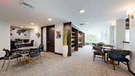 Located at 4351 Little Egret Rd in Kissimmee, Florida, this single-floor communal hallway and lounge area offers a thoughtfully designed environment that balances functionality with comfort. The space is finished with neutral-toned carpet tiles throughout, complemented by clean white walls and dark wood accents that lend a polished, contemporary aesthetic.

A central feature of the space is a built-in dark wood shelving unit that serves as a partial divider, creating distinct zones within the open layout. One side accommodates a business lounge area equipped with a workstation, computer terminal, and a round table with task chairs — ideal for productivity. A decorative world map accent on the wall adds a global design touch to this working nook.

Transitioning through the space, a bright lounge alcove benefits from expansive floor-to-ceiling windows that frame lush Florida greenery and palm trees, flooding the area with natural light. Woven lounge chairs and a round bistro table create an inviting setting for relaxation or casual conversation. Decorative circular wall art and coastal-themed photography panels add artistic character throughout.

The extended hallway corridor is wide and well-lit with recessed lighting, flanked by large multi-pane windows and accented with tall potted plants and a bench seating nook — creating a welcoming, resort-inspired passage through the building.