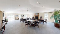 Located at 1420 Southern Ridge Lane in Charlottesville, Virginia, this well-appointed single-floor community space offers a generous and versatile layout designed for gathering and social activity. The main area is anchored by a striking floor-to-ceiling stone fireplace with a warm wood mantel, providing a welcoming focal point for the lounge seating area, which includes plush sofas and armchairs arranged for comfortable conversation. Multiple round tables with leather rolling chairs offer additional seating for group activities and dining. Vaulted ceilings with a ceiling fan lend an airy, open feel to the space, while recessed lighting ensures a bright, inviting atmosphere throughout. Numerous windows and French doors flood the interior with natural light and offer views of the surrounding neighborhood. A fully equipped kitchen runs along one side of the space, featuring rich wood cabinetry, dark granite countertops, stainless steel appliances including a refrigerator, range, microwave, and dishwasher, and light tile flooring that contrasts beautifully with the carpeted main area. A bookshelf and wall-mounted television provide entertainment options, rounding out this thoughtfully designed community hub.