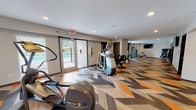 This fitness center is designed to accommodate a wide range of workout needs across multiple interconnected zones. The most visually striking element throughout the main cardio corridor is the bold herringbone-patterned flooring, incorporating shades of orange, gray, brown, and natural wood tones that lend a modern, energetic aesthetic to the space.

The primary cardio area is generously sized and lined with Precor equipment, including ellipticals, treadmills, and a rowing machine. Recessed lighting illuminates the room evenly, and a glass door with sidelights provides direct access to an outdoor pool deck, allowing natural light to filter into the space. A wall-mounted television is positioned for convenient viewing during workouts.

A dedicated free weights and functional training room features carpeted flooring, a dumbbell rack, medicine balls, exercise benches, and yoga mats, offering ample open floor space for stretching and bodyweight exercises. A teal accent wall adds a pop of color, and large mirrors enhance the sense of depth. Wall-mounted televisions are present in this area as well.

A connecting corridor with the same herringbone flooring leads to accessible restrooms, maintaining a cohesive design language throughout. The overall ambiance is contemporary, functional, and well-maintained.