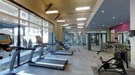 This modern fitness center offers a well-appointed, single-floor layout designed to support a full range of workout routines. The space is anchored by a row of premium cardio machines, including TRUE-brand treadmills and ellipticals, positioned along expansive floor-to-ceiling windows that invite abundant natural light and provide views of the surrounding building exterior. The flooring throughout is durable, low-pile carpet tile suited for both cardio and strength activities. Overhead, a combination of suspended rectangular pendant fixtures and recessed lighting ensures the entire area is thoroughly illuminated. Exposed concrete ceiling elements and structural columns lend an industrial character to the space, contrasting with the sleek, modern equipment. Toward the rear, a glass-enclosed exercise studio provides a dedicated area for group classes or personal training, outfitted with additional equipment and mirrored walls that enhance the sense of openness. A strength training zone occupies the far end of the main floor, featuring cable machines and free weights. The overall ambiance is clean, functional, and energizing, making this fitness center a comprehensive facility for residents or building occupants seeking a professional-grade workout environment.