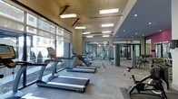 This modern fitness center offers a well-appointed, single-floor layout designed to support a full range of workout routines. The space is anchored by a row of premium cardio machines, including TRUE-brand treadmills and ellipticals, positioned along expansive floor-to-ceiling windows that invite abundant natural light and provide views of the surrounding building exterior. The flooring throughout is durable, low-pile carpet tile suited for both cardio and strength activities. Overhead, a combination of suspended rectangular pendant fixtures and recessed lighting ensures the entire area is thoroughly illuminated. Exposed concrete ceiling elements and structural columns lend an industrial character to the space, contrasting with the sleek, modern equipment. Toward the rear, a glass-enclosed exercise studio provides a dedicated area for group classes or personal training, outfitted with additional equipment and mirrored walls that enhance the sense of openness. A strength training zone occupies the far end of the main floor, featuring cable machines and free weights. The overall ambiance is clean, functional, and energizing, making this fitness center a comprehensive facility for residents or building occupants seeking a professional-grade workout environment.