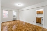 This single-floor residence is defined by its warm, classic character and thoughtful layout. The main living area is spacious and open, showcasing beautifully polished parquet hardwood flooring arranged in a distinctive geometric pattern that adds visual richness underfoot. Neutral cream-toned walls keep the space feeling bright and inviting, while natural light filters in through double windows equipped with an in-wall air conditioning unit. A radiator beneath the windows provides efficient heating. The living area connects seamlessly to a separate kitchen, accessible through a doorway framed by clean white trim. The kitchen is outfitted with warm-toned wood cabinetry, a granite-style countertop with an integrated sink, a gas range, and a full-size refrigerator — offering a complete and functional cooking space. An arched doorway leads to additional areas of the residence, adding an architectural detail that softens the overall aesthetic. The full bathroom features a clean, all-white palette with a combined tub and shower, a toilet, a vanity with granite countertop, and speckled tile flooring. Throughout the residence, consistent white trim and baseboards provide a crisp, cohesive finish that ties each space together cohesively.