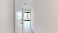 This efficiently designed one-bedroom, one-bathroom unit offers a well-organized layout across a single floor. The open-concept living and kitchen area serves as the heart of the space, where floor-to-ceiling windows frame sweeping city skyline views, flooding the interior with natural light. Exposed concrete ceilings add an industrial character, complemented by warm light wood flooring that runs throughout the unit.

The kitchen is thoughtfully appointed with light wood cabinetry extending to the ceiling, stainless steel appliances including a full-size refrigerator, dishwasher, and range, along with a black matte faucet over a recessed sink. Track lighting above the kitchen provides practical illumination.

A hallway connects the living area to the private bedroom, which maintains the same clean aesthetic with white walls and wood flooring. A dedicated closet with a full-width metal hanging rod offers organized storage. A sliding frosted glass door near the entrance adds a sleek, space-conscious design element.

The bathroom features a glass-enclosed shower with white tile surround, a floating wood-toned vanity with a white countertop, a framed mirror, matte black fixtures, and terrazzo-style flooring, completing the unit's modern, cohesive design.