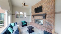 This single-level lounge offers a well-appointed outdoor living experience defined by its architectural character and thoughtful amenities. The focal point of the space is a full-height brick fireplace wall, constructed with warm-toned brick and finished with a rustic wood mantel. A flat-screen television is mounted above the fireplace, making the area ideal for both casual relaxation and social gatherings.

The lounge features soaring ceilings that enhance the sense of openness, complemented by a modern ceiling fan with natural wood-toned blades that provides airflow throughout the space. A row of arched windows along one wall introduces natural light and adds an elegant architectural detail to the otherwise clean, white-painted walls.

The flooring consists of neutral stone tile laid in a varied pattern, adding texture and visual interest underfoot. Seating includes modular wicker furniture with vibrant teal cushions and striped accent pillows, creating a lively yet comfortable atmosphere. A wicker coffee table anchors the seating arrangement. The overall ambiance is relaxed yet refined, blending indoor comfort with an open-air aesthetic.
