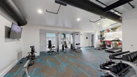 This fitness center is a thoughtfully designed workout facility that offers a comprehensive range of exercise equipment across a single open floor. The space is anchored by a striking, bold geometric accent wall featuring a vibrant mix of red, yellow, teal, and neutral tones, adding an energetic and modern aesthetic to the environment. Exposed black ductwork runs overhead against a clean white ceiling, lending an industrial-chic character to the interior. Recessed lighting throughout ensures the space is well-lit, while multiple windows allow natural light to filter in.

The cardio area features multiple treadmills and a rowing machine, positioned to face a wall-mounted flat-screen television for an engaging workout experience. The strength training zone includes a multi-station cable machine and a squat rack with a bench, catering to a full range of resistance exercises. A dedicated free weight station houses a tiered dumbbell rack stocked with a variety of weights, complemented by medicine balls and additional accessories nearby. The flooring throughout consists of a distinctive teal-striped carpet tile, combining durability with visual appeal. An on-site restroom adds convenience, and direct exterior access via a glass door connects the fitness center to the broader property.