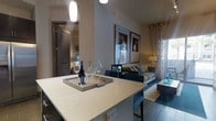 This thoughtfully designed single-floor residence showcases a seamless open-concept layout that connects the kitchen and living areas into one cohesive, functional space. The kitchen is anchored by a generous island with light stone countertops, offering both prep space and casual seating for bar-style dining. A pendant light fixture hangs above, providing focused illumination that complements the overall modern aesthetic.

The living area flows naturally from the kitchen, featuring a comfortable sofa accented with teal and neutral-toned throw pillows. A pair of framed art pieces mounted on the wall adds a curated, polished feel, while a warm table lamp introduces a soft ambiance to the space. Sheer curtains frame a window that invites natural light to fill the room, enhancing the open and airy atmosphere.

Dark tile flooring grounds the space and provides a striking contrast to the light countertops and neutral wall tones, lending a sophisticated, contemporary character throughout. A corridor visible from the kitchen suggests additional private rooms beyond, offering a sense of depth and separation between living and personal spaces. Overall, the residence presents a clean, modern design with carefully chosen finishes and furnishings that balance style and everyday comfort.