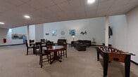 Located at 901 Draper Lofts in Schenectady, NY, this generously sized single-floor community amenity space offers a versatile and well-appointed environment for residents to gather, relax, and socialize. The main lounge area is immediately impressive, featuring soaring ceilings that lend a distinctly open and airy character to the space. Neutral-toned carpet, crisp white walls, and tasteful decorative artwork — including teal-accented abstract pieces and wall clocks — create a welcoming, contemporary aesthetic throughout. The lounge is furnished with a sectional sofa, armchairs, and side tables arranged around a wall-mounted flat-screen television, providing a comfortable setting for casual gatherings. A foosball table and high-top bistro tables offer additional recreational and social options. The open layout flows naturally into a kitchenette area equipped with cabinetry, a refrigerator, and a microwave, ideal for hosting events or informal get-togethers. A dedicated restroom with accessible fixtures is also available within the space. A secondary area features additional high-top seating, drop ceilings, and double doors for convenient access. Throughout, recessed and ceiling-mounted lighting ensures the space is well-lit and functional at all times.