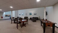 Located at 901 Draper Lofts in Schenectady, NY, this generously sized single-floor community amenity space offers a versatile and well-appointed environment for residents to gather, relax, and socialize. The main lounge area is immediately impressive, featuring soaring ceilings that lend a distinctly open and airy character to the space. Neutral-toned carpet, crisp white walls, and tasteful decorative artwork — including teal-accented abstract pieces and wall clocks — create a welcoming, contemporary aesthetic throughout. The lounge is furnished with a sectional sofa, armchairs, and side tables arranged around a wall-mounted flat-screen television, providing a comfortable setting for casual gatherings. A foosball table and high-top bistro tables offer additional recreational and social options. The open layout flows naturally into a kitchenette area equipped with cabinetry, a refrigerator, and a microwave, ideal for hosting events or informal get-togethers. A dedicated restroom with accessible fixtures is also available within the space. A secondary area features additional high-top seating, drop ceilings, and double doors for convenient access. Throughout, recessed and ceiling-mounted lighting ensures the space is well-lit and functional at all times.