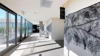This single-floor corridor serves as a refined transitional space within a larger building, offering access to key amenities including a Fitness Center, Clubhouse, and Elevators, as indicated by sleek overhead directional signage. The hallway is characterized by its contemporary design, with clean white walls and large-format light-toned flooring that reflects abundant natural light streaming in through floor-to-ceiling glazed panels lining one side. These expansive windows create a bright, open atmosphere while visually connecting the interior to the outdoors. A standout feature of the corridor is its bold, large-scale abstract wall murals rendered in fine black linework against the white walls, adding a strong artistic identity to the space. A minimalist white armchair is positioned mid-corridor, offering a subtle seating moment. Concrete columns punctuate the hallway at intervals, contributing structural rhythm without interrupting the flow. The overall aesthetic is modern and curated, balancing functional wayfinding with thoughtful artistic elements to create an engaging and visually cohesive environment.