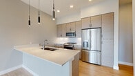 This contemporary single-floor residence showcases a thoughtfully designed open-concept layout with high-end finishes and a clean, modern aesthetic. The kitchen serves as the centerpiece of the living space, featuring light wood-grain flat-front cabinetry with matte black hardware, crisp white quartz countertops, and a large island with a double undermount sink. Stainless steel appliances — including a gas range, built-in microwave, and French door refrigerator — elevate the kitchen's functionality, while sleek cylindrical pendant lights hang from soaring ceilings to enhance the contemporary ambiance. Light hardwood flooring runs throughout the main living and kitchen areas, contributing to a bright and airy atmosphere. The bedrooms are finished with plush neutral carpeting and feature large windows and sliding glass doors that open to a balcony, offering expansive outdoor views of the surrounding neighborhood and skyline. One bedroom includes an elegant tray ceiling with recessed lighting, adding architectural depth. The bathroom is well-appointed with marble-look tile flooring, a wood-tone vanity with a quartz countertop, a dark-framed mirror, and a full tub-shower combo with white subway tile surround and chrome fixtures. Overall, the space exudes a refined, modern character with consistent attention to detail.
