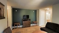 This single-story space presents a welcoming living area defined by a striking deep green accent wall that anchors the room and adds a rich, earthy character to the interior. The wall serves as a natural focal point, framing a wooden dresser used as a media console with a flat-screen television mounted above it. Two small, round wooden side tables flank the dresser symmetrically, contributing to a balanced and functional layout.

The flooring throughout is warm-toned hardwood-style planking, lending the space a natural, cohesive feel that complements the green wall and wooden furnishings. A textured ceiling adds subtle visual interest overhead, while clean white baseboards and trim provide a crisp contrast to the bolder design elements.

A white paneled door is visible toward the rear of the room, suggesting additional spaces beyond. The layout feels open and practical, with enough room for comfortable seating and movement. Overall, the space achieves a grounded, casual ambiance through its earthy color palette, natural materials, and straightforward design—making it a functional and visually cohesive living environment.