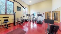 Located at 130 Rollins Ave in Rockville, Maryland, this fitness center offers a well-equipped, single-floor exercise facility designed to support a range of workout routines. The space features a bold red rubberized floor that adds both durability and energy to the environment, complemented by warm yellow walls and ample natural light streaming in through multiple large windows that offer views of surrounding greenery.

The cardio area is furnished with several Life Fitness treadmills and elliptical machines, providing users with a variety of aerobic options. The strength training section includes a multi-station cable machine, a lat pulldown and row machine, adjustable benches, free weights, and additional resistance equipment, making it suitable for both beginner and experienced fitness enthusiasts. A wall-mounted flat-screen television is positioned for viewing during workouts.

Full-length mirrors along one wall enhance the sense of openness and allow users to monitor their form. Instructional fitness posters are displayed throughout the room for guidance. The facility also includes an adjoining restroom accessible directly from the main exercise floor, adding convenience for users. The overall layout is open and functional, offering enough room to move between stations comfortably while maintaining a practical, purpose-driven atmosphere.