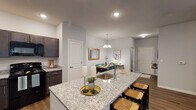 Located at 7405 Fegenbush Ln in Louisville, KY, this single-floor residence offers a well-designed, open-concept layout that maximizes both functionality and comfort. The kitchen is a standout feature, showcasing rich dark cabinetry, granite countertops, recessed lighting, and a full suite of black appliances including a refrigerator, range, and microwave. A granite-topped island with bar seating serves as the centerpiece, seamlessly connecting the kitchen to the adjacent dining and living areas. The dining area is defined by warm wood-look flooring and an elegant chandelier overhead, creating an inviting space for gatherings. The living room offers a relaxed, open feel with a ceiling fan, recessed lighting, and a door providing access to an exterior patio or entry. Two well-appointed bedrooms feature plush carpeting and generous windows that allow for natural light. The primary bedroom includes wall-mounted sconce lighting and access to a private bathroom. A second full bathroom features a granite-topped vanity with dark cabinetry and a tub-shower combination. Throughout the residence, neutral gray walls, wood-look vinyl flooring, and clean architectural lines contribute to a cohesive, modern aesthetic that is both practical and stylish.
