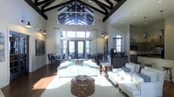 This impressive single-floor space is defined by its dramatic architectural centerpiece: a soaring vaulted ceiling adorned with dark exposed wood beams arranged in a striking geometric pattern. A large clerestory window at the apex of the ceiling floods the interior with natural light, creating a bright and airy atmosphere throughout the day. The main living area is elegantly appointed with crisp white upholstered sofas and lounge chairs arranged around a tufted brown leather ottoman, all set atop a light-toned area rug over rich dark hardwood floors. Tall, floor-to-ceiling French doors and large windows line the walls, offering views of the outdoor surroundings while further enhancing the sense of openness. Soft white curtains frame the windows, adding a refined touch to the space. A dining area is visible toward the rear, furnished with dark chairs and tables suited for gathering. An arched doorway leads to additional areas, including what appears to be a service or refreshment counter, adding functional versatility to the space. Wall-mounted sconces and recessed lighting complement the natural illumination, ensuring a warm and welcoming ambiance at any hour. The overall design balances grand architectural details with clean, contemporary furnishings.