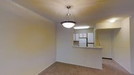 This single-floor space presents a functional and straightforward layout, with the dining area and kitchen connected through a convenient pass-through counter. The dining area features neutral-toned carpeting and warm beige walls, with crown molding along the ceiling adding a subtle decorative touch. A polished pendant light with a bowl-shaped frosted glass shade hangs prominently, serving as a central focal point for the dining space. The pass-through counter creates an open connection to the kitchen, which is equipped with white cabinetry arranged in a practical configuration, a white refrigerator, and a fluorescent ceiling light providing ample task lighting. The white cabinetry, combined with the light walls, gives the kitchen a clean, bright appearance. A standard entry door is visible adjacent to the kitchen, framed neatly within the layout. The overall ambiance of the space is modest and functional, with consistent neutral tones throughout that provide a versatile backdrop. White baseboards frame the carpeted floor, contributing to a cohesive and tidy finish throughout the space.