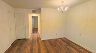 This single-floor residence offers a clean, straightforward layout characterized by rich, dark hardwood flooring that flows seamlessly throughout the main living areas and central hallway. The dining area is highlighted by an elegant candelabra-style chandelier that adds a classic touch to the otherwise neutral, understated space. White walls provide a bright, airy backdrop throughout, complemented by crisp white baseboards that frame the hardwood floors with a polished finish. A central hallway extends from the main living area, connecting the space to additional rooms beyond, creating a practical and intuitive flow. Overhead lighting in the hallway ensures the passage remains well-lit and welcoming. A louvered closet door near the dining area provides convenient storage access while maintaining the clean aesthetic of the space. Ceiling vents indicate a central HVAC system for year-round climate control. The overall ambiance is warm and inviting, with the combination of natural wood tones and soft white walls creating a neutral canvas well-suited for a variety of interior styles.