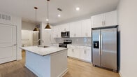 Located on Populus Drive in Pooler, Georgia, this single-floor home delivers a clean, modern aesthetic with thoughtfully selected finishes throughout. Light-toned luxury vinyl plank flooring flows seamlessly through the main living areas, complemented by crisp white walls and abundant recessed lighting.

The kitchen serves as a focal point of the home, showcasing white shaker-style cabinetry extending to the ceiling for generous storage, granite countertops, a subway tile backsplash, and a full suite of stainless steel appliances including a range, over-the-range microwave, and a French door refrigerator with an ice and water dispenser. A central island with a granite top and an undermount sink is accented by two striking gold-toned pendant lights, creating an ideal space for both meal preparation and casual dining.

The open living area features a ceiling fan, recessed lighting, and a window overlooking a green outdoor view, filling the space with natural light. A half-wall divider elegantly defines the transition between living spaces while maintaining an open feel.

The bathroom is finished with matching white cabinetry, granite countertops, gold hardware accents, and a tiled tub-shower surround. The bedroom features plush carpet flooring and provides access to a walk-in closet with built-in shelving. The overall ambiance is fresh, bright, and move-in ready.