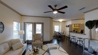 This single-floor residence offers an open-concept layout that seamlessly connects the living, dining, and kitchen areas into one cohesive and functional space. The living area is anchored by rich hardwood flooring and features a ceiling fan with integrated lighting, complemented by crown molding that adds a refined architectural detail throughout. Natural light flows generously into the space through multiple windows, including a set of French doors that open onto a private balcony, creating a bright and airy atmosphere. The living area provides ample room for comfortable seating arrangements, while the dining area accommodates a full dining table and chairs, situated just steps away from the kitchen. The kitchen is partially open to the main living space via a raised bar counter with barstool seating, making it ideal for casual dining and entertaining. Warm, neutral tones on the walls harmonize with the dark hardwood floors to create an inviting and cohesive aesthetic. The crown molding, ceiling details, and consistent flooring throughout contribute to a polished and well-appointed interior that balances both style and practicality.