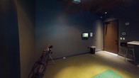 This dedicated indoor golf simulator room is thoughtfully designed to deliver an immersive golfing experience in a controlled environment. The space is finished with deep charcoal-toned walls and a dark ceiling, creating a focused, distraction-free atmosphere that enhances the visual impact of the simulation system.

At the heart of the room is a High Definition Golf simulator, featuring a large projection screen framed by black curtain surrounds that span the full width of one wall. The ceiling-mounted projector delivers crisp, high-resolution imagery of world-class golf courses, transporting players to realistic virtual environments. A secondary wall-mounted monitor is positioned to the side, providing additional game data and scoring information during play.

The flooring consists of a dual-tone artificial turf hitting mat — a green strip laid over a broader yellow-green surface — providing a realistic feel underfoot for each shot. A golf bag with clubs stands ready near the hitting area, and golf balls are conveniently staged on the floor.

A bistro-style table with bar stools is positioned near the rear of the room, offering a comfortable spot for spectators or players to relax between rounds. The overall ambiance is intimate and purpose-built, making this simulator room an ideal venue for practice, entertainment, or competitive play year-round.