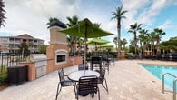 The outdoor amenity area at 980 Courthouse Rd presents a well-appointed single-level courtyard space designed for both relaxation and social gatherings. The patio is finished with attractive paver stonework that extends throughout the space, providing a clean and durable surface underfoot.

A standout architectural feature is the stucco-clad outdoor fireplace, framed with classic detailing and topped with a substantial chimney. Adjacent to the fireplace is a built-in grilling station, making this area well-suited for outdoor cooking and entertaining. The combination creates a focal point that anchors the entire courtyard.

Multiple round patio tables, each accompanied by sleek black metal chairs, are arranged across the space. Vibrant green market umbrellas provide shade and add a pop of color against the warm tones of the paver surface. The open-air setting is framed by tall, mature palm trees that lend a tropical ambiance and a sense of established landscaping.

A black wrought-iron perimeter fence defines the boundary of the space while maintaining visibility to the surrounding grounds. The courtyard benefits from generous natural light and open skies, creating an airy, resort-like atmosphere ideal for outdoor gatherings throughout the day.