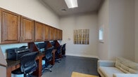This single-floor space functions as a versatile business center combining a well-equipped computer lab with a welcoming lounge area. The layout is divided into two distinct zones that together create a balanced environment suited for both focused work and casual interaction.

The workstation area is anchored by a row of built-in dark wood desks, each featuring individual privacy dividers and equipped with desktop computers and shared printing capabilities. Rich, warm-toned cabinetry stretches across the full length of the wall above the desks, providing generous storage while contributing a polished, professional aesthetic. Ergonomic task chairs accompany each station, and a skylight overhead supplements the recessed lighting to keep the workspace well-illuminated.

The lounge zone near the entrance offers a more relaxed atmosphere, furnished with neutral-toned upholstered armchairs arranged around a wooden side table and accented by a woven area rug. Large gridded windows dressed with floor-length charcoal drapes allow natural light to filter in while framing outdoor views. The entry features double glass-paneled doors with keycard access, reinforcing the space's secure and semi-private character.

Throughout, the polished concrete flooring and neutral gray walls create a clean, contemporary backdrop, while carefully chosen artwork adds warmth and visual interest to the overall ambiance.