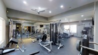 This fitness center offers a well-organized and fully equipped space designed to accommodate a variety of workout routines. The interior is finished with neutral gray walls and a smooth, dark rubberized flooring that provides both durability and comfort underfoot. Recessed lighting throughout the ceiling ensures the space is consistently well-lit, while multiple ceiling fans promote air circulation during intense workouts.

Large mirrored panels line one wall, visually expanding the space and allowing users to monitor their form during exercise. The equipment selection is extensive, including a multi-station cable and weight machine positioned centrally, a leg press machine, and an assortment of additional strength-training equipment visible in the mirrored reflection. Along the perimeter, cardio machines such as treadmills and elliptical trainers are arranged to maximize floor space and usability.

A window on the far wall introduces natural light, adding to the overall brightness and openness of the room. The layout is thoughtfully arranged to allow users to move freely between equipment, making the fitness center both functional and accessible.