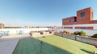 This single-story residence offers a well-appointed interior complemented by an impressive rooftop outdoor space. The entryway greets residents with rich, dark hardwood-style flooring and neutral-toned walls, establishing a clean and modern aesthetic carried throughout the unit. A security panel near the entry door adds a practical touch.

The open-concept living and dining area is defined by a seamless transition between hardwood-style flooring and plush carpeting, delineating functional zones within the shared space. Crown molding and decorative column accents lend architectural character, while ceiling fans contribute to the overall comfort of the space. A pendant light fixture highlights the dining area, and granite countertops on the adjacent kitchen bar provide both visual appeal and utility.

Beyond the interior, the rooftop terrace serves as a notable feature, offering a generous outdoor area with artificial turf, multiple raised wooden planting beds for gardening, and open views of the surrounding cityscape. A small seating area with teal chairs adds a welcoming touch to the rooftop retreat. The terrace is bordered by low white parapet walls and a neighboring brick structure, creating a semi-enclosed yet open-air environment ideal for relaxation or urban gardening.