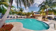 This single-story property showcases a spectacular outdoor living area at its heart — a generously sized freeform swimming pool with crystal-clear turquoise water, edged by elegant travertine pavers that extend into a broad, sun-drenched deck. Towering palm trees frame the pool on all sides, creating a lush, resort-like canopy that offers both shade and a sense of tropical seclusion. The pool area is well-appointed with lounge chairs and colorful tropical plantings, adding vibrant splashes of red and green throughout the landscape. A charming pool house anchors the rear of the outdoor space, designed in a Mediterranean-inspired style with warm stucco walls, a classic terracotta tile roof, and arched entryways. The pool house features large upper-level windows that allow for natural light and views over the surrounding landscape. A perimeter wall encloses the grounds, enhancing privacy while maintaining the serene, self-contained atmosphere. The overall ambiance of this property is that of a private tropical retreat, combining lush natural landscaping with thoughtfully designed architectural elements to create an inviting and relaxing outdoor environment suited for warm-weather living.