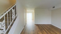 This two-story residence offers a well-organized layout designed for comfortable everyday living. The main level is anchored by a bright, open hallway that connects the key areas of the home, finished with rich hardwood flooring that adds warmth and continuity throughout the space. Natural light streams in through vertical blinds covering a large window or sliding door at the end of the hallway, creating an airy and inviting atmosphere. The walls are finished in crisp white, complementing the light-toned textured ceiling and contributing to the clean, neutral aesthetic of the interior. A staircase with a decorative white metal railing leads to the upper level, adding a subtle ornamental touch to the entryway. Doorways along the hallway provide access to additional rooms, suggesting a thoughtful floor plan that balances open circulation with defined private spaces. The overall ambiance is bright, clean, and move-in ready, with a classic residential character. The hardwood floors and neutral palette create a versatile backdrop suitable for a variety of interior styles. The second floor likely offers additional bedrooms or living spaces, completing the home's multi-level functionality.