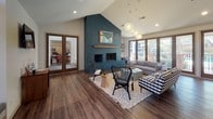 This single-floor space is defined by its striking architectural character, most notably the vaulted ceilings that rise dramatically throughout, creating an expansive, airy atmosphere. Rich hardwood-style flooring runs continuously across the interior, unifying the open layout and complementing the warm, eclectic aesthetic.

The main lounge area centers around a floor-to-ceiling teal painted brick fireplace fitted with a wood mantle and built-in shelving, serving as the room's most commanding focal point. A large sectional sofa, rattan chairs, and patterned textiles furnish this area, while a wall of sliding glass doors floods the space with natural light and opens directly to an outdoor patio. A unique hanging egg chair adds a playful design element nearby.

An adjacent seating nook features a vaulted ceiling peak with an arched window and built-in bench below, accompanied by a wall-mounted television. Pendant globe lights hang at varying heights throughout, adding warmth and visual interest.

A central dark-topped island anchors a bar or wet area with white cabinetry and black countertops, suitable for entertaining. A glimpse into an adjoining room reveals fitness equipment, indicating a dedicated exercise space within the facility.