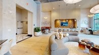 Located at 3558 Elm St in Atlanta, GA, this single-floor lobby and lounge space offers a generously sized, thoughtfully designed environment that blends contemporary elegance with warm, residential comfort. The open-plan layout is anchored by soaring ceilings adorned with a reflective mirrored tile ceiling and oversized cloud-like pendant light fixtures that create a dramatic yet welcoming atmosphere. Floor-to-ceiling black-framed glass windows and doors flood the interior with natural light while offering views of the surrounding streetscape. The entry corridor features striking terrazzo flooring and curated wall art, setting a refined tone from the moment of arrival. The main lounge area centers around a sculptural curved sectional sofa in a warm neutral tone, complemented by accent chairs and layered decorative pillows. A dedicated seating alcove showcases a rich amber-toned banquette with textured wall paneling, an artistic relief sculpture, and statement pendant lighting. A bar or service counter area with a warm mosaic tile backdrop and integrated display screen adds a functional yet stylish focal point. A cozy secondary lounge nook features golden upholstered seating with indirect LED cove lighting along a soft-toned tile wall. Lush potted plants throughout add a refreshing natural element, completing this polished and inviting space.