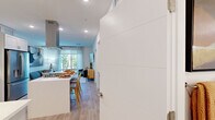This thoughtfully designed one-bedroom, one-bathroom residence makes excellent use of its compact single-floor layout, offering a bright and modern living experience. The entrance opens directly into a sleek, open-concept kitchen that serves as the heart of the home. The kitchen is outfitted with white and light gray shaker-style cabinetry extending to the ceiling, a striking stainless steel island range hood, stainless steel appliances, and a center island with a crisp white countertop and bar seating — ideal for casual dining and entertaining. Light wood-tone flooring flows throughout, lending warmth and continuity to the space. A dedicated display ledge with additional cabinetry adds functional storage and a stylish focal point. The bedroom is a tranquil retreat, featuring large floor-to-ceiling windows that frame lush, tree-lined outdoor views and allow generous natural light. The bathroom is finished with light wood-look tile flooring, gray shaker vanity cabinetry with a white countertop, a large mirror, and a full soaking tub surrounded by elegant marble-style tile surround. Recessed lighting throughout the home contributes to a clean, polished ambiance. A dedicated closet and entrance area complete this efficiently designed, contemporary residence.