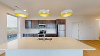 This contemporary single-floor residence is thoughtfully designed with a clean, modern aesthetic throughout. The open-concept kitchen serves as a central highlight, showcasing rich dark-brown cabinetry, a classic white subway tile backsplash, and a full suite of stainless steel appliances including a refrigerator, electric range, and built-in microwave. A generous island with a light quartz countertop and an undermount sink anchors the space, illuminated overhead by a trio of striking white-and-gold dome pendant lights that add a warm, modern flair. Light wood-look flooring flows through the main living areas, contributing to a bright and airy atmosphere. The living area features a sliding glass door that opens to a private balcony, offering pleasant views of the surrounding neighborhood and treeline. The bedrooms are finished with plush carpeting and feature ample natural light through well-placed windows. A spacious walk-in closet with wire shelving provides generous storage, while a dedicated in-unit laundry area adds everyday convenience. The bathroom is finished in crisp white with a shower/tub combination, a large vanity mirror, and clean, simple fixtures. Throughout the unit, neutral tones and modern finishes create a cohesive, move-in-ready environment.