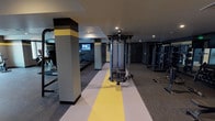 This fitness center presents a thoughtfully designed workout facility with a contemporary aesthetic defined by dark charcoal walls, bold yellow accent stripes, and durable rubber flooring. A distinctive yellow and white striped pathway runs through the center of the space, visually dividing the floor into dedicated workout zones while adding a dynamic design element.

The cardio area is positioned near large windows that allow natural light to enter and provide views of the surrounding exterior, creating a more open and energizing atmosphere for exercise. This section houses multiple treadmills, elliptical machines, stationary bikes, and a rowing machine, along with a wall-mounted fan and television for added comfort during workouts.

The strength training area occupies a generous portion of the space and features a multi-station cable machine as a central fixture, complemented by a power rack, plate-loaded machines, a dumbbell rack, and various benches. Structural columns accented with yellow bands are integrated throughout, maintaining the facility's cohesive color scheme. Recessed lighting illuminates the entire space, and a water station near the entrance ensures easy access to hydration. Mirrors along the walls enhance the sense of depth throughout.