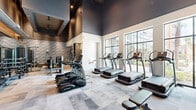 Located at 2180 Pickens Street in Columbia, SC, this contemporary fitness center delivers a premium workout experience within a thoughtfully designed, single-floor space. The facility is accessed through striking floor-to-ceiling black-framed glass doors, setting the tone for the modern aesthetic found throughout.

The main cardio area is anchored by an impressive row of True-brand treadmills, stair climbers, and ellipticals, all positioned to take full advantage of the expansive, multi-pane windows that flood the space with natural light and offer views of the surrounding greenery. Soaring ceilings with dark upper accents add a dramatic architectural dimension and a sense of openness.

A dedicated strength and functional training zone features a full array of True-brand selectorized machines, a multi-station cable system, and TRX and XRT suspension training stations mounted against a warm wood-panel accent wall. Full-length mirrors amplify the space and allow for proper form monitoring. A well-stocked dumbbell rack lines the wall nearby.

The flooring throughout features a sophisticated marble-look tile, complementing the layered design palette of dark ceilings, textured stone accent walls, and warm wood tones. Exposed ductwork in the strength area adds an industrial touch. The result is a cohesive, high-end fitness environment designed for both functionality and visual appeal.