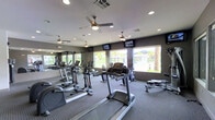 This single-story community clubhouse is designed with a contemporary aesthetic that blends functionality and sophistication. The main lounge area immediately impresses with its soaring coffered ceiling, anchored by a large, sculptural floral chandelier that serves as a striking focal point. Light hardwood-style flooring extends throughout, complementing the neutral color palette. Floor-to-ceiling windows dressed in sheer white drapery flood the space with abundant natural light while framing views of the surrounding landscape. A sleek bar counter runs along one side of the lounge, lined with modern chrome and metal barstools, making it ideal for social gatherings. Comfortable lounge seating is arranged near the windows, creating an inviting area to relax. An accent wall featuring a stylized mural adds an artistic touch to the overall design.

Adjacent to the lounge, the fitness center is well-appointed with a comprehensive selection of cardio equipment, including treadmills, ellipticals, and a rowing machine. Multiple ceiling fans maintain air circulation, while wall-mounted televisions provide entertainment. Large windows offer views of palm trees and the surrounding community, creating a bright and motivating workout environment.